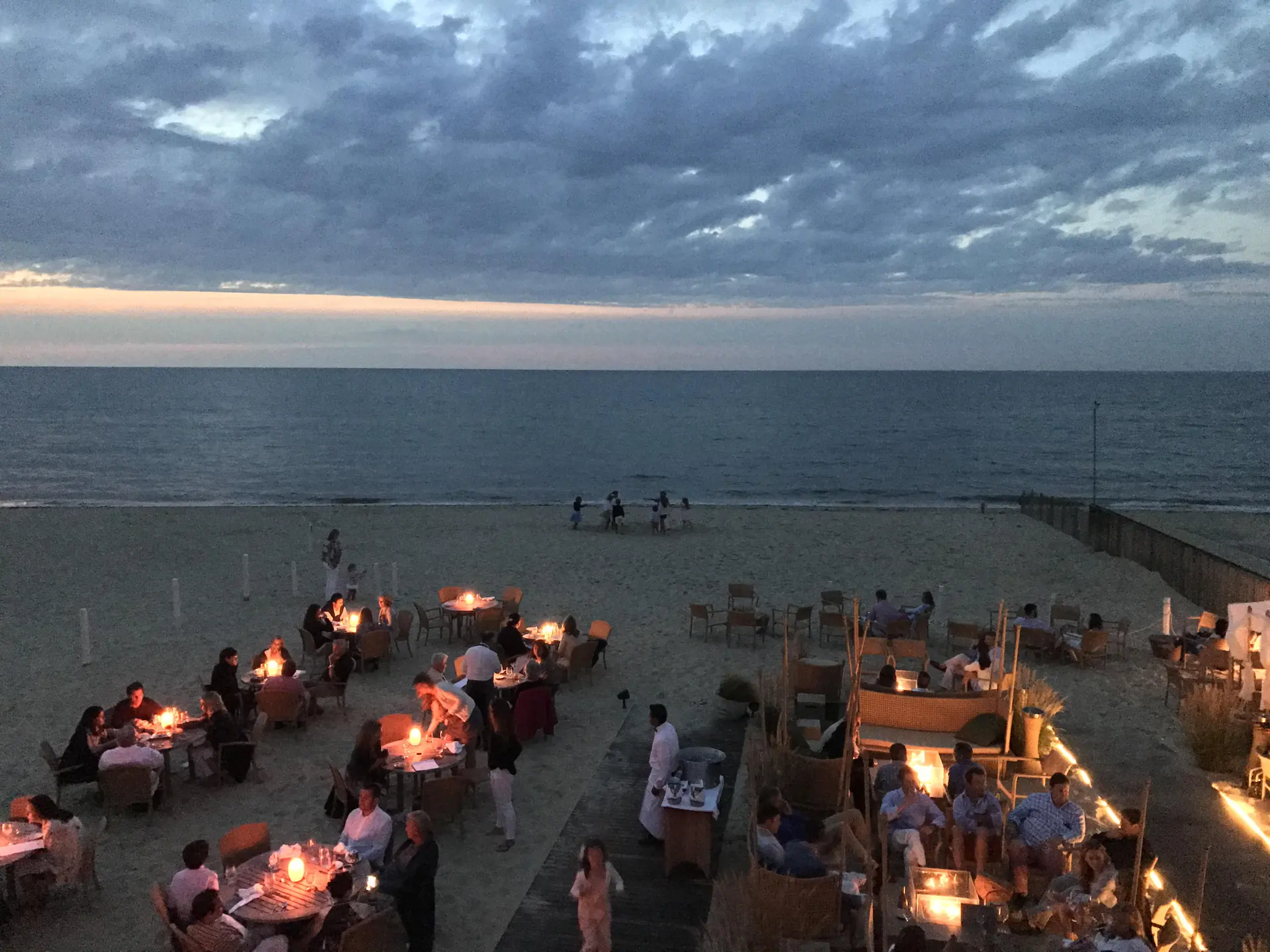 Candlelit dinner tables on the sand at dusk on Nantucket's north shore — the summer evening the island's rental market sells