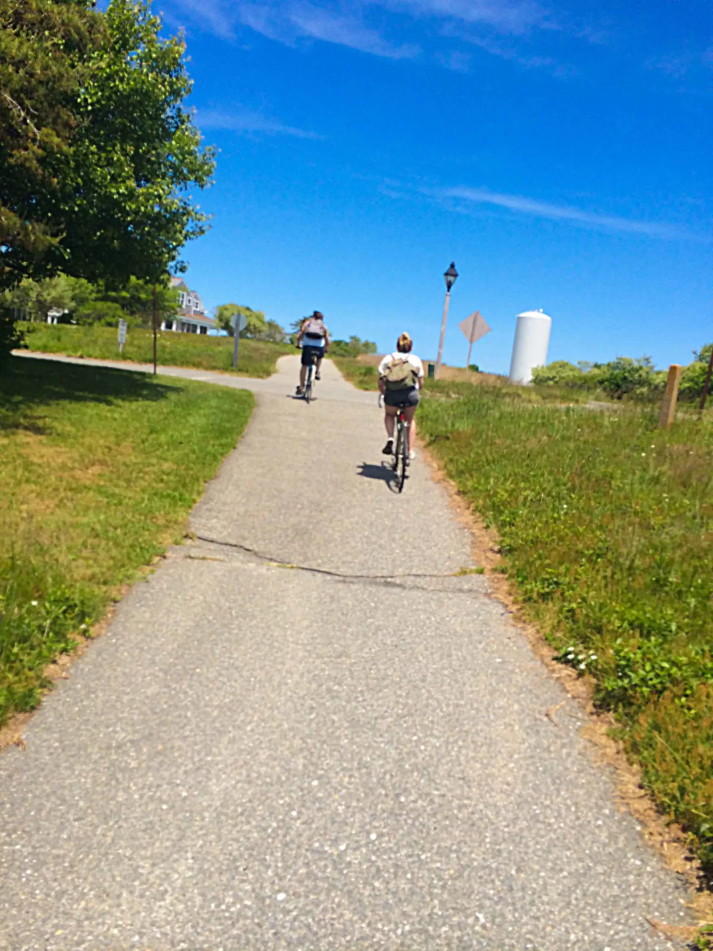 Two cyclists riding away up a paved Nantucket sidepath under a blue sky, grass verges either side, a water tower and lamppost ahead on the right