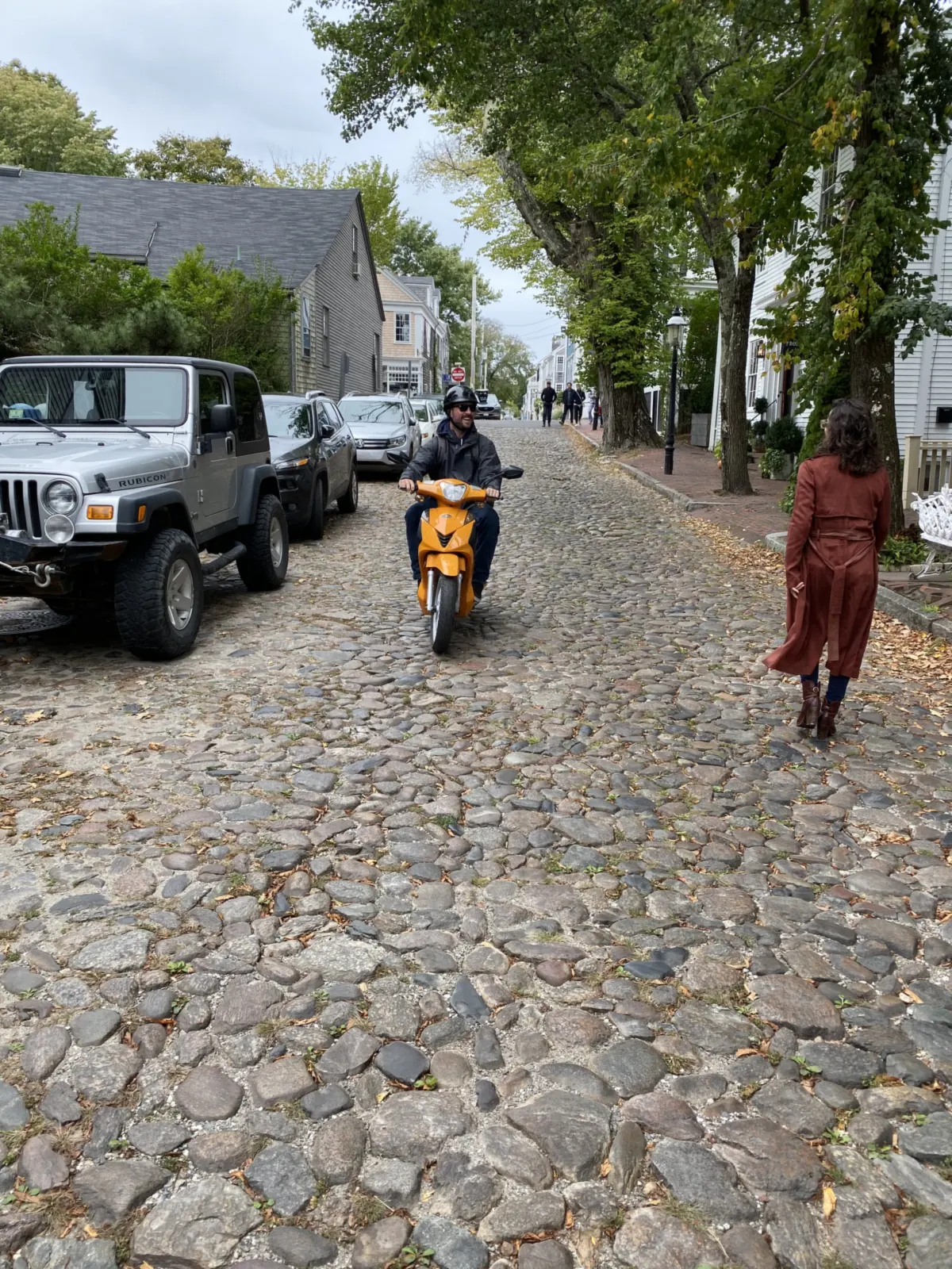 A moped rider on a cobblestone street in downtown Nantucket, with a parked Jeep on the left and a pedestrian on the brick sidewalk under the trees