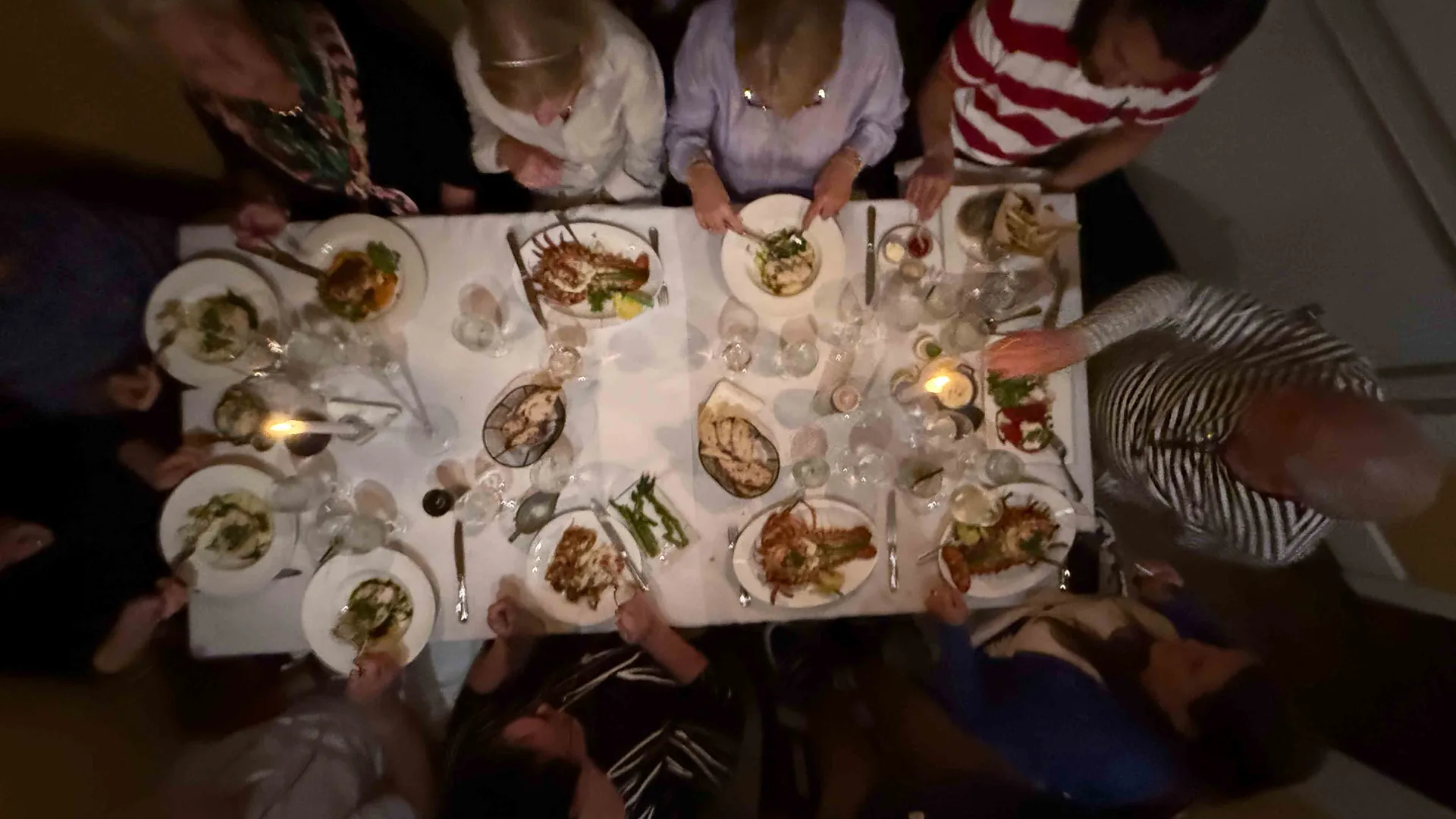 A long candlelit dinner table seen from above, lobster and island plates passing between friends