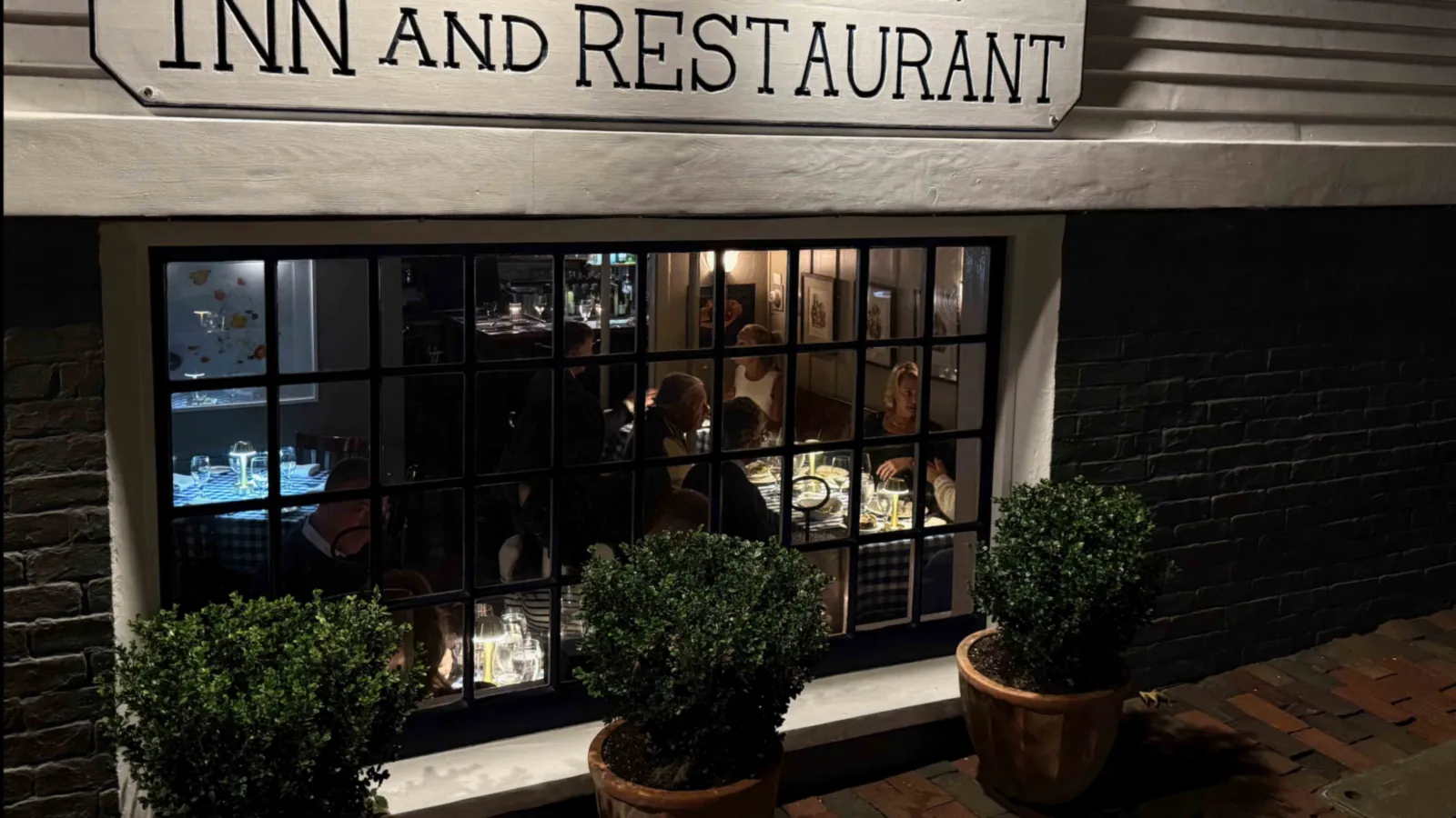A small-paned restaurant window glowing at night on a downtown lane, diners at checked tablecloths inside