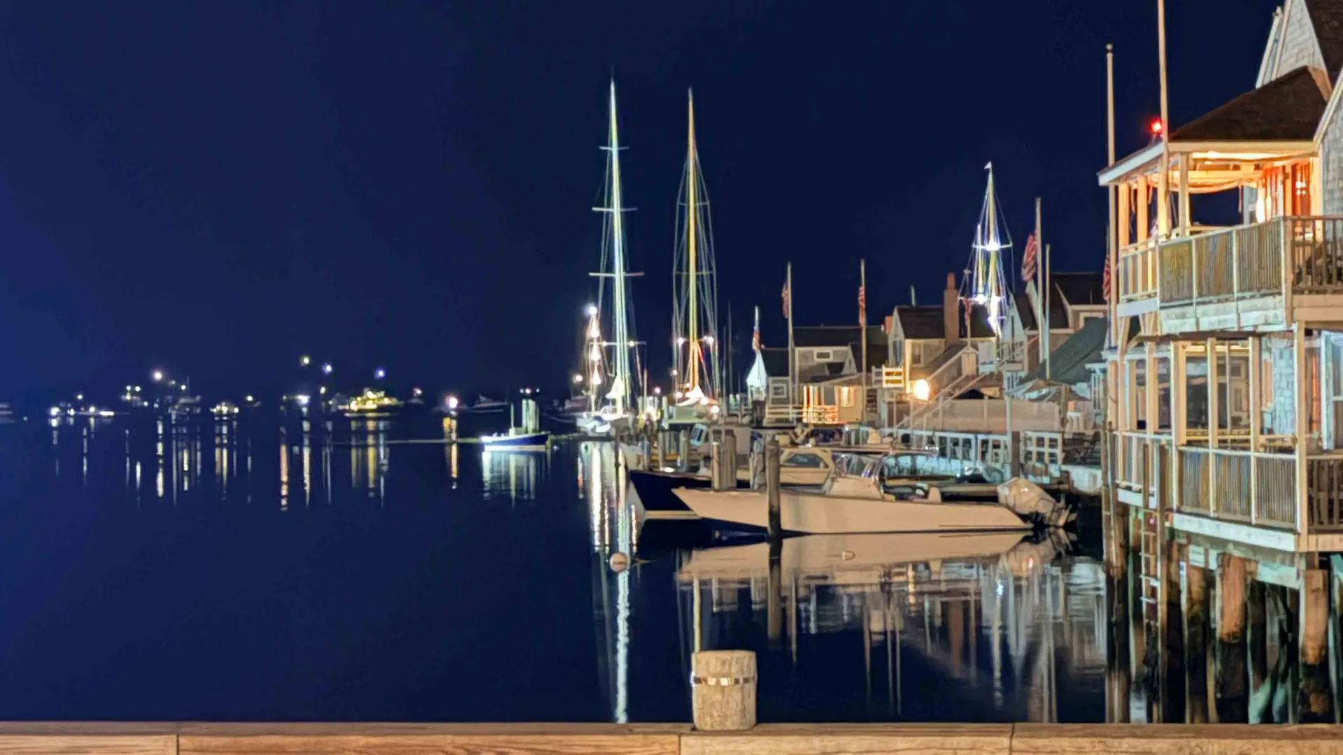 Sailboats moored for the night at Nantucket's Easy Street basin, tall masts strung with lights against a dark sky, the harbor town's shingled waterfront buildings glowing beside their reflections in still water