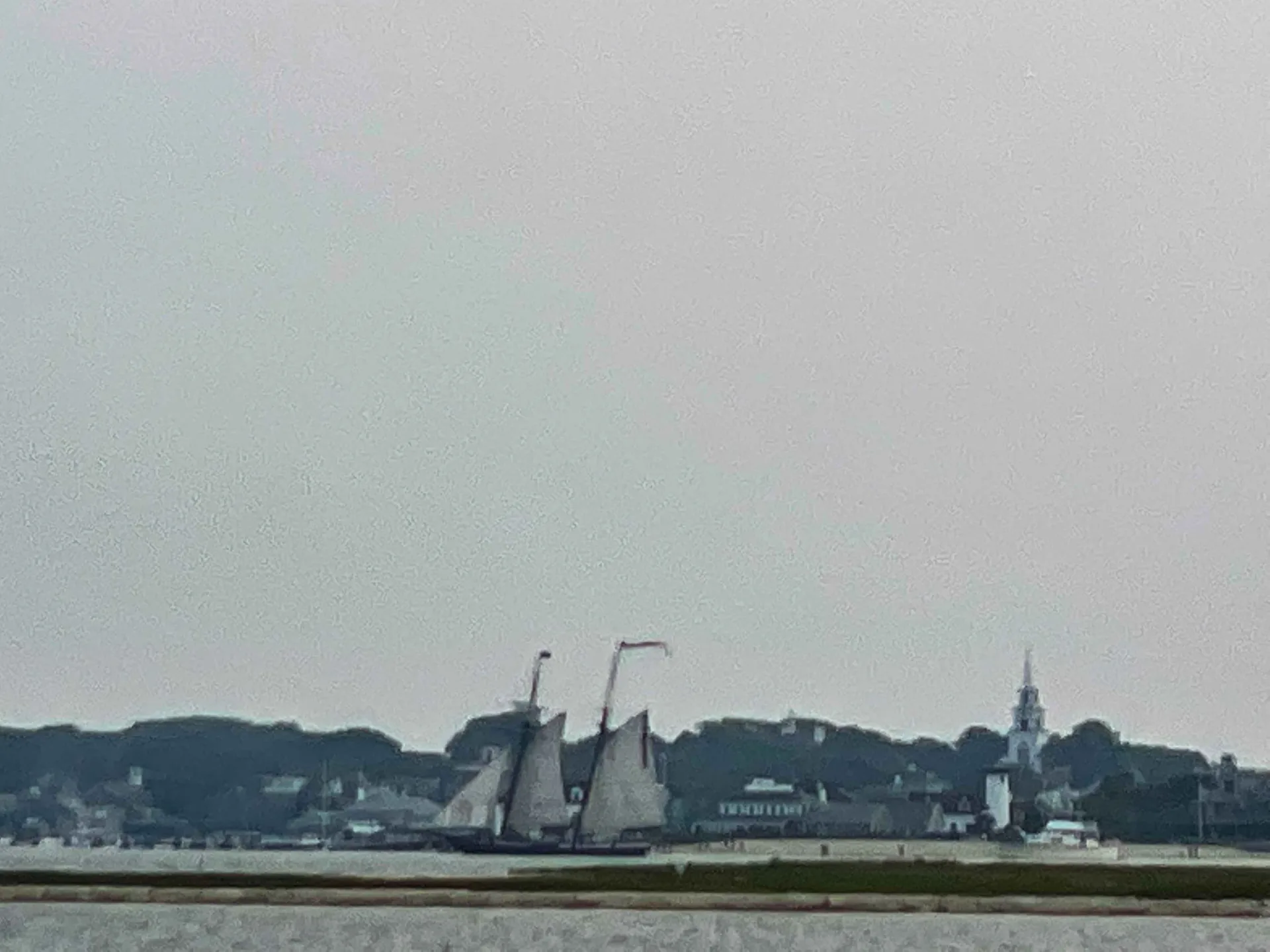 A schooner under full sail off Nantucket town on a grey morning, the church steeple and the shingled waterfront behind it