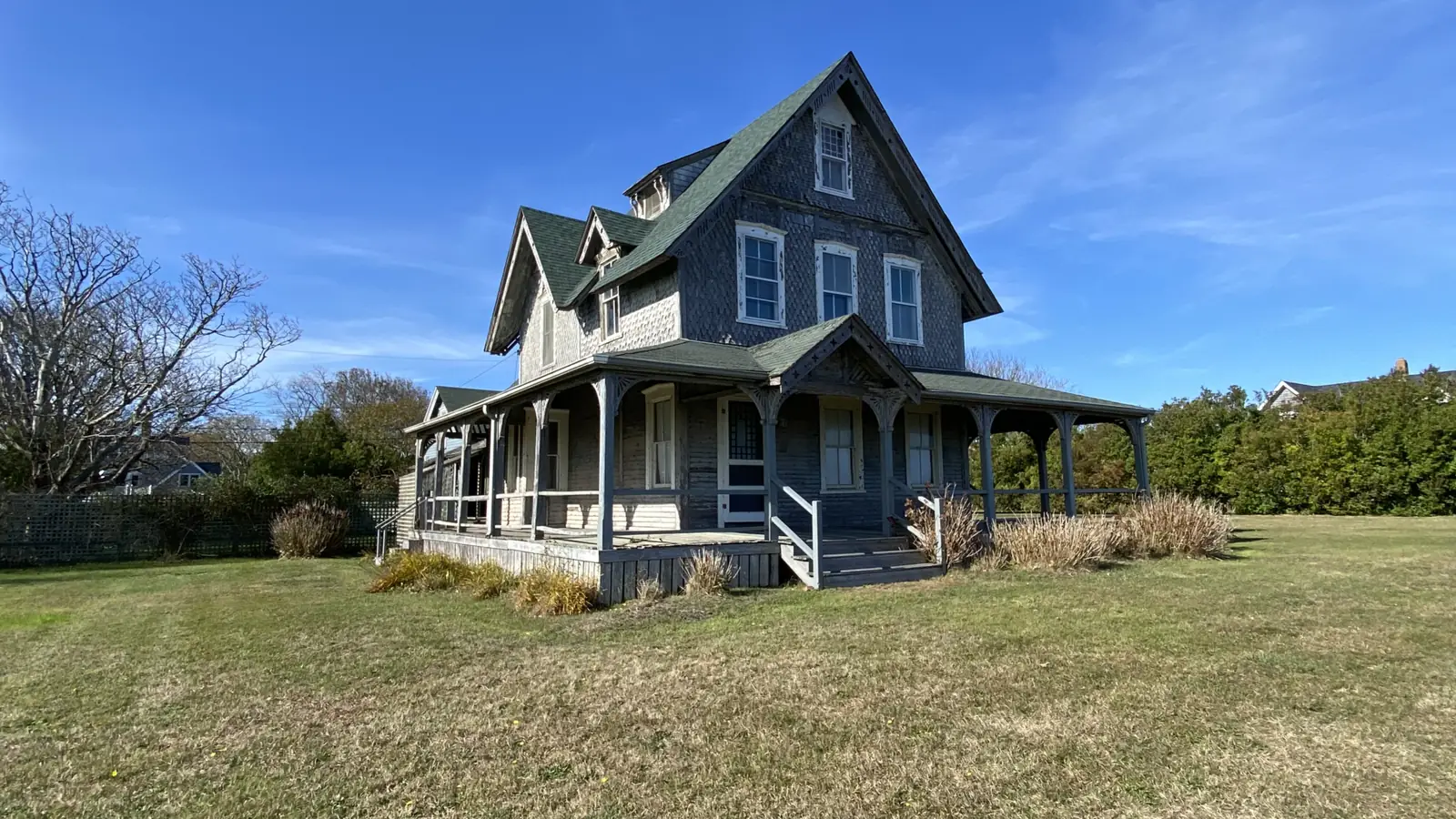A gray-shingled Nantucket cottage with a deep wraparound porch on an open lawn, bare trees behind it on a clear November afternoon