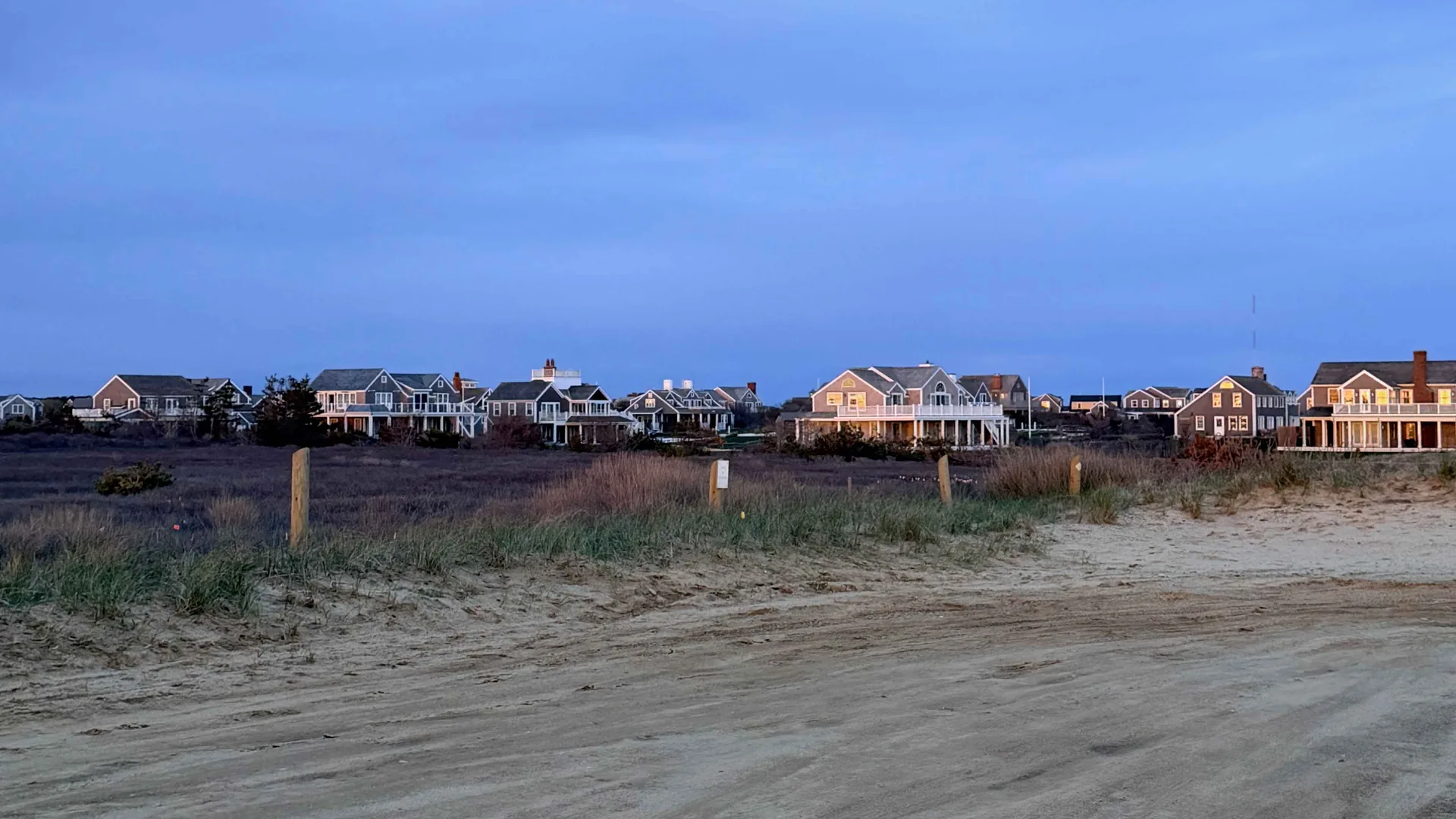 A row of gray-shingled Nantucket houses with lit windows and white-railed decks at blue hour, seen across beach-grass dunes and a sand road under a deep evening sky