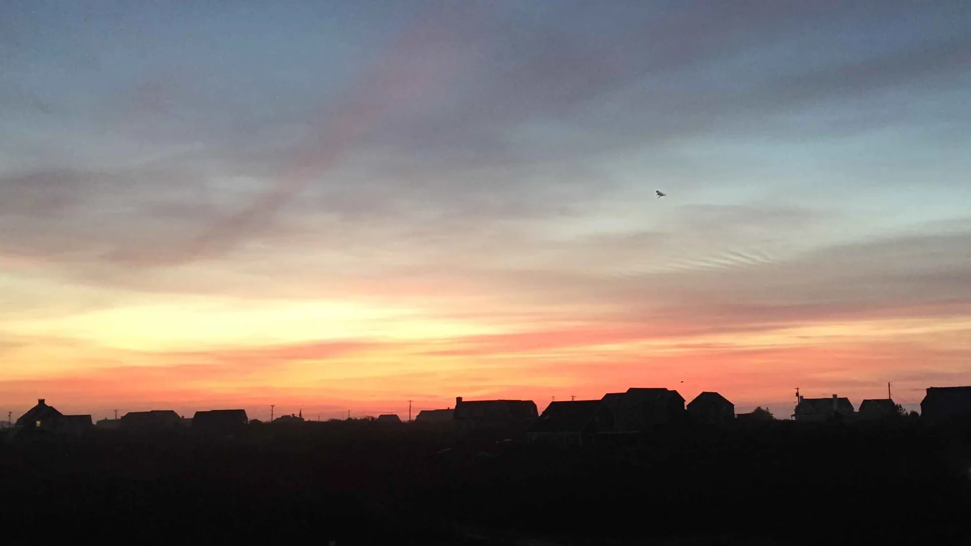 Sunrise over Tom Nevers, Nantucket: silhouetted rooflines along the horizon under a blazing orange-to-pink dawn sky, a small plane crossing high above