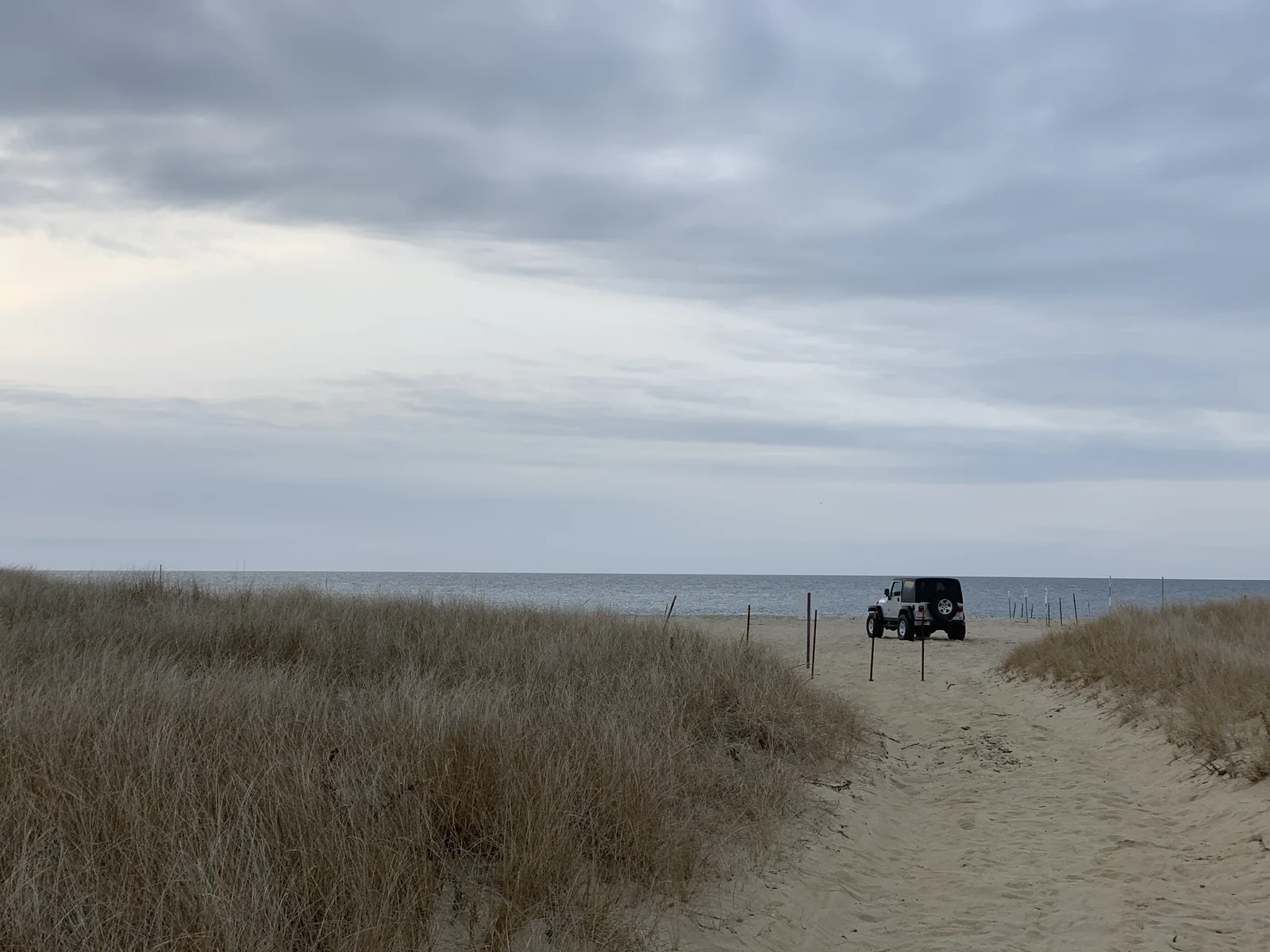 A winter beach path through pale dune grass on Nantucket under a heavy gray sky, a four-wheel-drive parked alone at the water