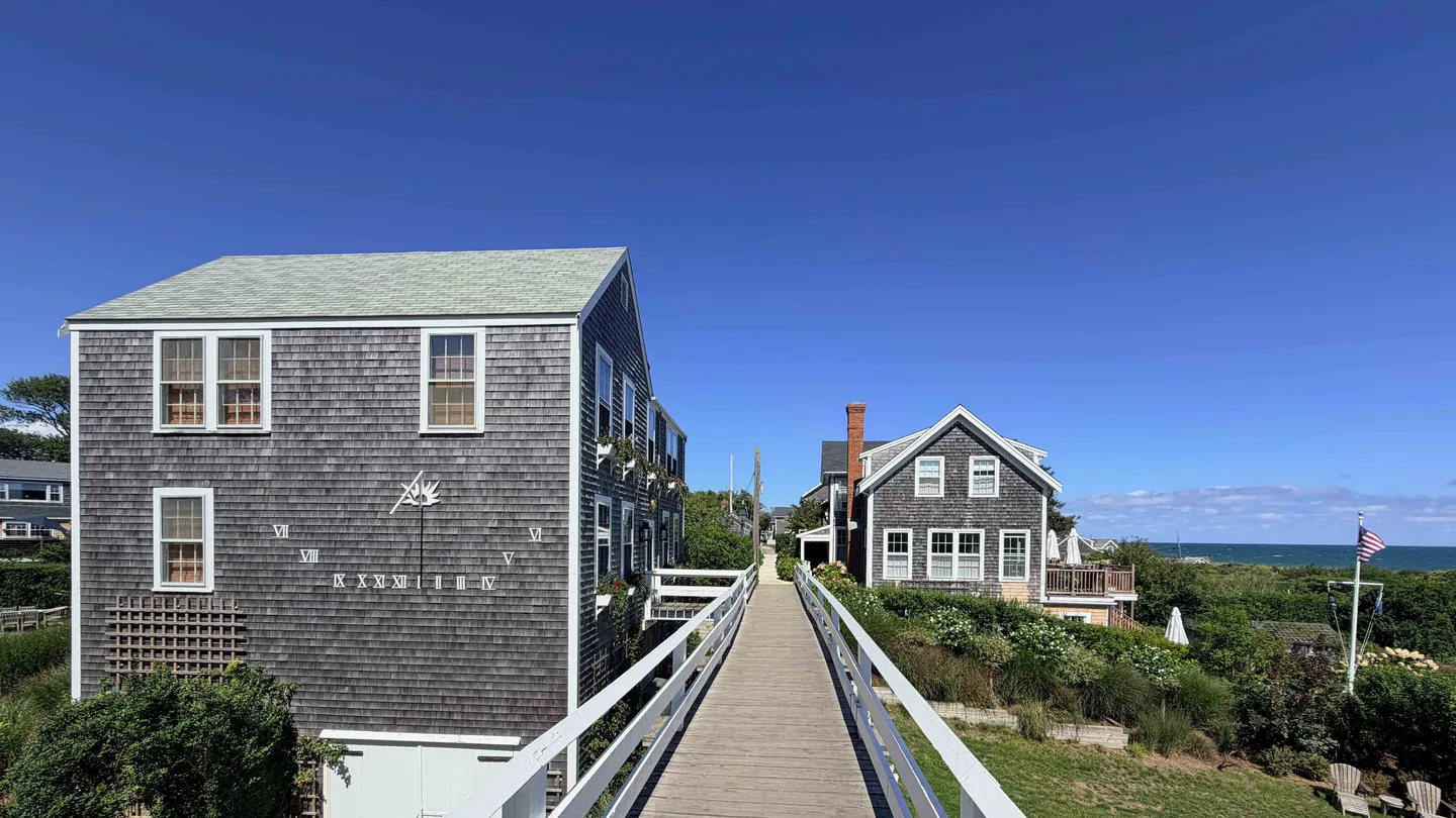 A wooden boardwalk between weathered shingled cottages in 'Sconset, Nantucket, one painted with a sundial, leading toward the Atlantic