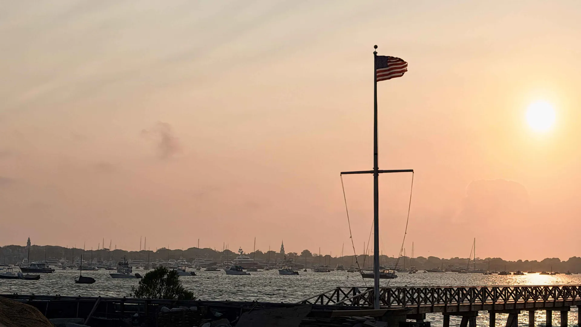 Nantucket harbor at sunset seen from Shimmo, with the town skyline behind a flagpole and moored boats